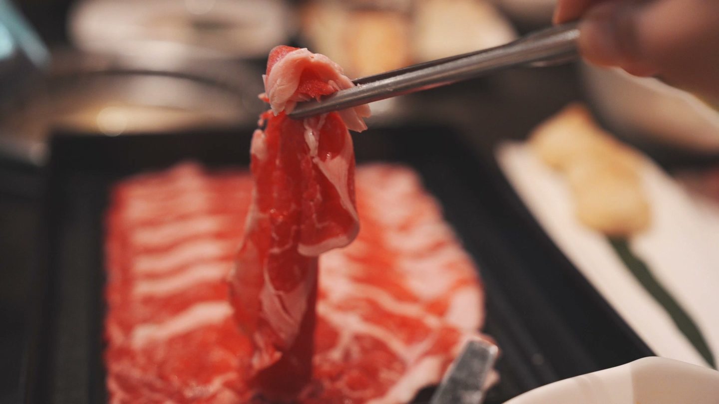 Chopsticks lifting a thin slice of marbled beef over a tray of sliced meat