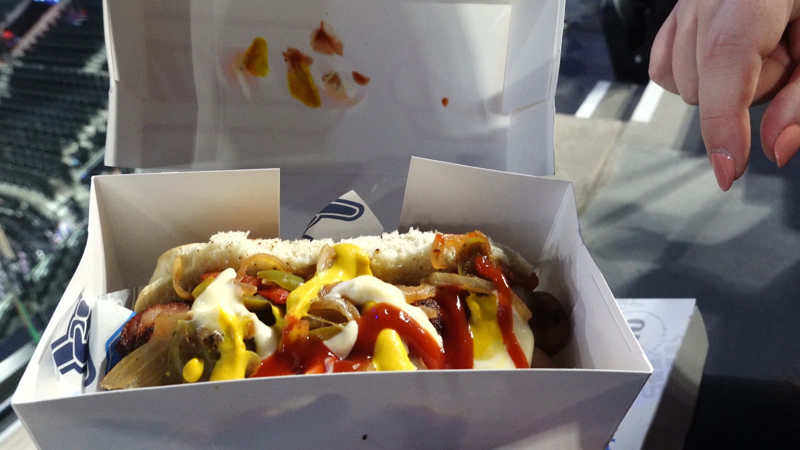 Bacon-wrapped LA street dog loaded with grilled onions, ketchup, mustard, mayo, and jalapeños in a box at Intuit Dome